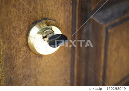 Close-up of a worn key inserted into an old, yellowed wooden door, highlighting a vintage keyhole and weathered knob. The rustic textures and faded colors emphasize age 120298049