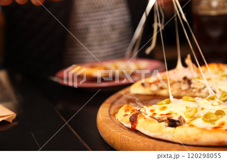 A woman enjoying a slice of pizza outdoors, with melted mozzarella cheese stretching from the slice. The pizza is topped with green olives 120298055