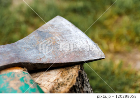 Close-up of an old, weathered hatchet embedded in a tree stump, with a blurred green background highlighting the rugged details of the axe. The aged metal and worn handle convey themes of rustic work Close-up of an old, weathered hatchet embedded in a tree stump, with a blurred green background highlighting the rugged details of the axe. The aged metal and worn handle convey themes of rustic work 120298058