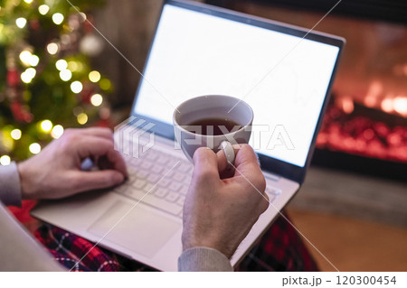 Christmas. Man with laptop sitting near christmas tree and fireplace with cup of tea. Concept of online service in holidays. Back view. Christmas. Man with laptop sitting near christmas tree and fireplace with cup of tea. Concept of online service in holidays. Back view. 120300454