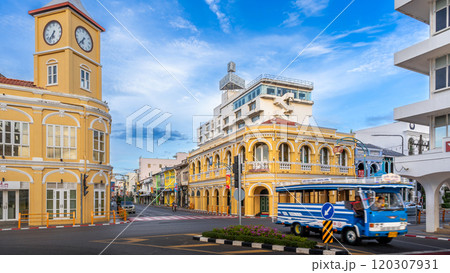 The iconic yellow clock tower and historic Chartered Bank building in Phuket Old Town, showcasing Sino-Portuguese architecture, frame a bustling intersection with a blue local bus passing by The iconic yellow clock tower and historic Chartered Bank building in Phuket Old Town, showcasing Sino-Portuguese architecture, frame a bustling intersection with a blue local bus passing by 120307931
