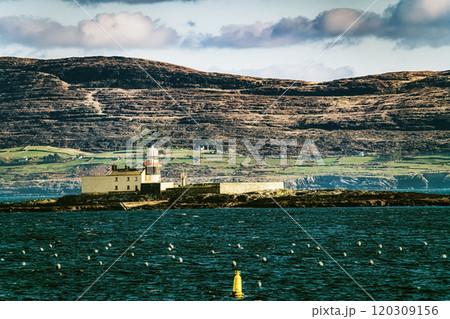 A lighthouse is positioned on a small island in a bay. The lighthouse is white with a red roof, and it is surrounded by dark gray rocks. The bay is mostly empty with a few small buoys in the water. A lighthouse is positioned on a small island in a bay. The lighthouse is white with a red roof, and it is surrounded by dark gray rocks. The bay is mostly empty with a few small buoys in the water. 120309156