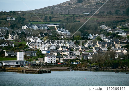 A picturesque scene of a small town nestled amidst a lush green hillside. The houses are mostly white and have black roofs, and there are trees and shrubs surrounding the area. 120309163