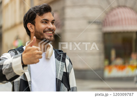 Happy young Indian man looking approvingly at camera showing thumbs up like gesture in city street Happy young Indian man looking approvingly at camera showing thumbs up like gesture in city street 120309787
