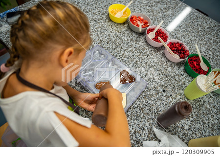 Little girl learning how to cook in a cooking class. Handmade dessert. Making chocolate candy, Child filling heart shaped mold with chocolate Little girl learning how to cook in a cooking class. Handmade dessert. Making chocolate candy, Child filling heart shaped mold with chocolate 120309905