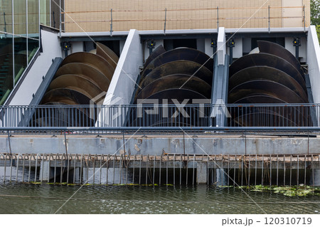 The image shows a set of large Archimedes screws used for water management or energy generation. The screws are housed within a structure beside a canal or river, with a metal railing in front. 120310719