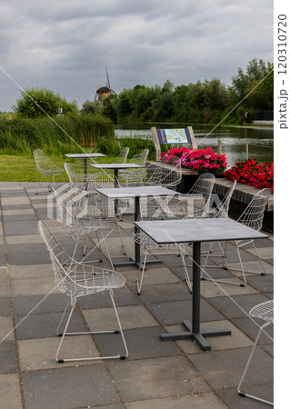 An outdoor cafe area with empty wire-frame chairs and tables on a stone patio next to a river. Red and pink flowers decorate the scene, and a windmill is visible in the background under a cloudy sky. 120310720