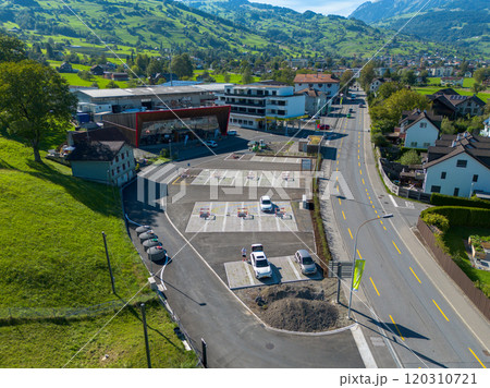 An aerial view of a small town showing a parking lot, a modern building, a curved road, and houses surrounded by green hills and mountains. The sky is clear, giving a vibrant, sunny atmosphere. 120310721