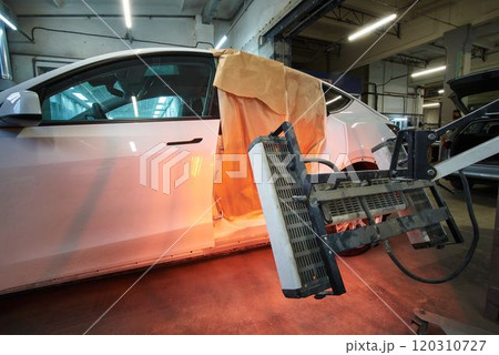 A white car is undergoing body repair with infrared drying equipment. The vehicle's side panel is masked with paper for protection during the heating process in an auto repair shop. 120310727
