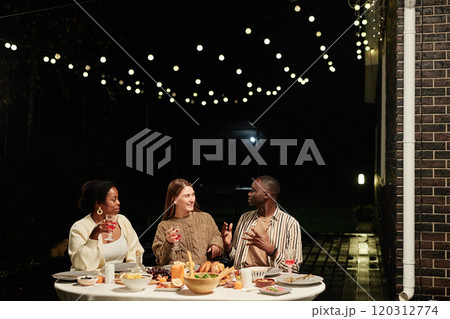 Group of three happy adult people enjoying dinner together outdoors at terrace decorated with twinkling lights, copy space 120312774