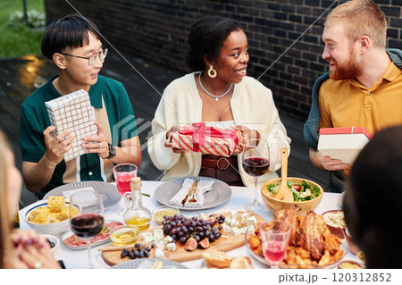 Vibrant portrait of smiling adult friends sharing gifts sitting at dinner table outdoors and celebrating holidays together 120312852