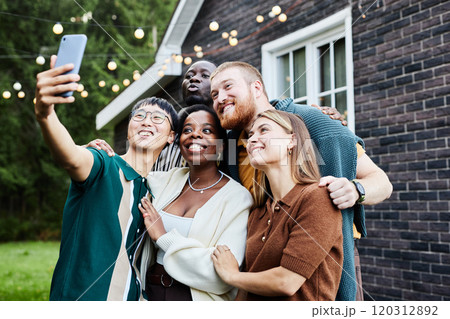Waist up portrait of multiethnic group of people taking selfie photo together outdoors by house decorated with fairy lights 120312892