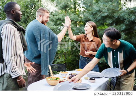 Side view at cheerful adult friends high five while setting dinner table together outdoors in green yard 120312900
