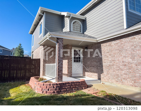 A suburban backyard during late fall, featuring a brick house with a cozy patio and a raised brick planter. The yard includes a green lawn with patches of fallen leaves, a concrete pathway, and a 120313098