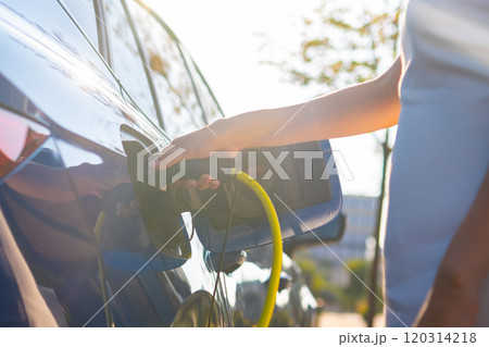 Close up woman hand connects a cable for charging to electric car against sunlight. Close up woman hand connects a cable for charging to electric car against sunlight. 120314218