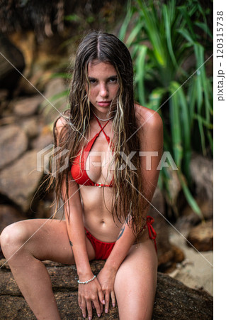 A Young Girl Dressed in a Bright Red Bikini Poses Cheerfully by a Scenic Waterfall 120315738