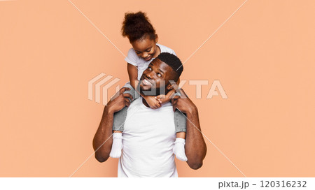 Happy African American father and his daughter posing together against a peach background. The father is smiling and looking to the side while holding his daughter on his shoulders 120316232