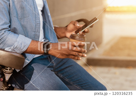 Modern Outdoor Leisure. Unrecognizable african man leaning on bicycle, using smartphone and drinking takeaway coffee outside Modern Outdoor Leisure. Unrecognizable african man leaning on bicycle, using smartphone and drinking takeaway coffee outside 120316235