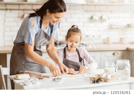 A mother and daughter are baking cookies together in their kitchen. The mother is guiding her daughter as they cut out shapes from cookie dough with cookie cutters. A mother and daughter are baking cookies together in their kitchen. The mother is guiding her daughter as they cut out shapes from cookie dough with cookie cutters. 120316261