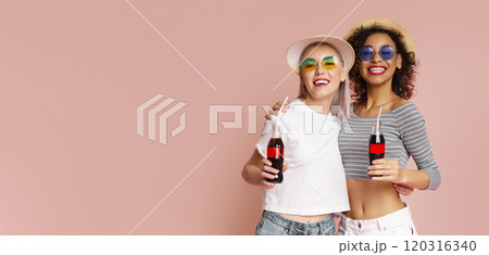 This image features two women in summer hats and sunglasses, smiling and holding soda bottles against a pink background. They are both wearing casual clothing, copy space 120316340