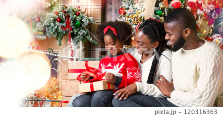 Family Christmas Concept. Cute little black girl opening gift box on Christmas eve while sitting on floor with parents in decorated living room 120316363