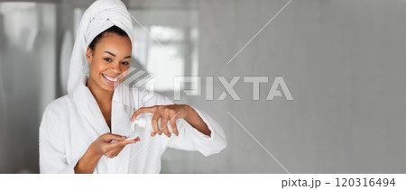 Black woman in a bathrobe and towel is happily applying a skincare product in her well-lit bathroom. Her morning routine highlights self-care and beauty practices with a relaxed atmosphere. 120316494