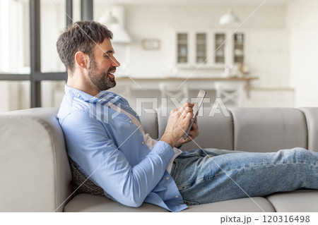 A man in a blue shirt and jeans sits comfortably on a gray couch, scrolling through a tablet. He leans back and smiles, looking relaxed and content. The room is bright and modern 120316498