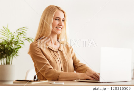 A woman with blonde hair smiles while working on a laptop at her home office desk. A woman with blonde hair smiles while working on a laptop at her home office desk. 120316509