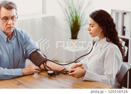 Hypertensive Crisis. Young latina nurse measuring blood pressure of elderly man at clinic office, copyspace Hypertensive Crisis. Young latina nurse measuring blood pressure of elderly man at clinic office, copyspace 120316526