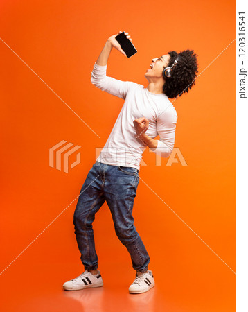 Young emotional black man singing his favorite song, using smartphone as microphone, orange studio background 120316541