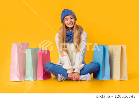 Shop as you can. Carefree blonde girl in winter clothes sitting on floor among shopping bags, pink studio background, copy space 120316545