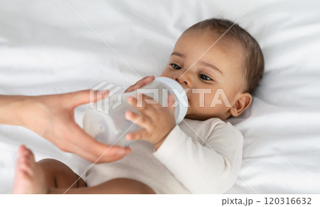 Childcare And Nutrition Concept. Portrait of cute little African American baby drinking milk from bottle, woman helping and holding, kid wearing bodysuit lying on the white blanket at home Childcare And Nutrition Concept. Portrait of cute little African American baby drinking milk from bottle, woman helping and holding, kid wearing bodysuit lying on the white blanket at home 120316632