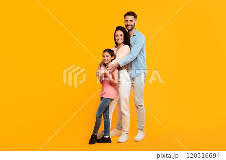 A family of three poses together against a bright yellow background. The father, mother, and young daughter are all smiling and looking at the camera 120316694