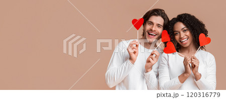 Love is in the air. Portrait of romantic multiracial couple holding red paper hearts on sticks and smiling at camera, posing over yellow background Love is in the air. Portrait of romantic multiracial couple holding red paper hearts on sticks and smiling at camera, posing over yellow background 120316779