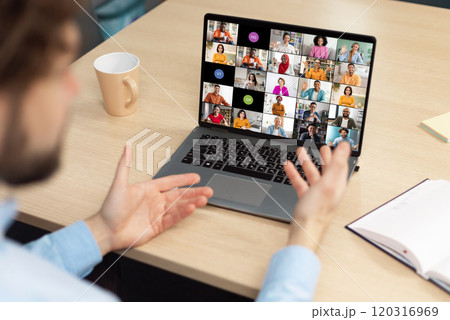 A man sits at a desk in front of a laptop, actively participating in a video conference meeting with colleagues. His hands are raised, gesturing and engaging with the online participants. 120316969