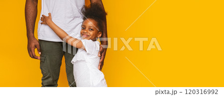 A cheerful young black girl with a big smile is affectionately hugging her father legs while standing on a vibrant yellow backdrop. The father stands calmly, showcasing a loving moment between them. A cheerful young black girl with a big smile is affectionately hugging her father legs while standing on a vibrant yellow backdrop. The father stands calmly, showcasing a loving moment between them. 120316992