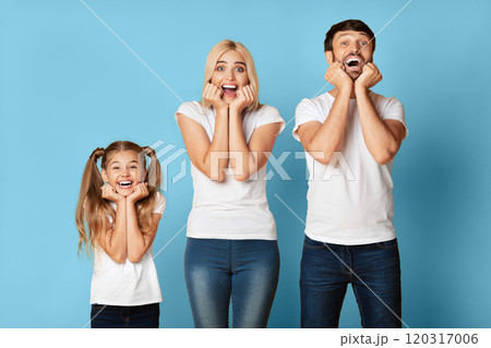 A young girl, her mother, and her father stand in front of a solid blue background. The family is smiling and looking up at the camera with their hands on their cheeks. 120317006