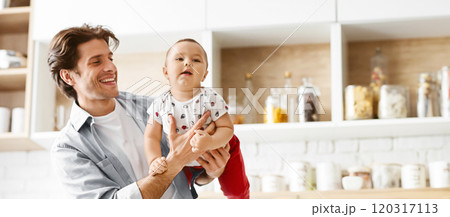 A smiling father holds his young son in the kitchen. The toddler is looking at the camera with a curious expression. The kitchen is modern and well-lit, with white cabinets and a tiled backsplash. A smiling father holds his young son in the kitchen. The toddler is looking at the camera with a curious expression. The kitchen is modern and well-lit, with white cabinets and a tiled backsplash. 120317113