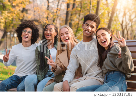 Positive international teenagers enjoying weekend at public park, sitting on bench and posing at camera Positive international teenagers enjoying weekend at public park, sitting on bench and posing at camera 120317193
