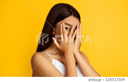A young Asian woman with long, dark hair covers her face with her hands in a gesture of shyness or embarrassment. She is standing in front of a bright yellow background A young Asian woman with long, dark hair covers her face with her hands in a gesture of shyness or embarrassment. She is standing in front of a bright yellow background 120317304
