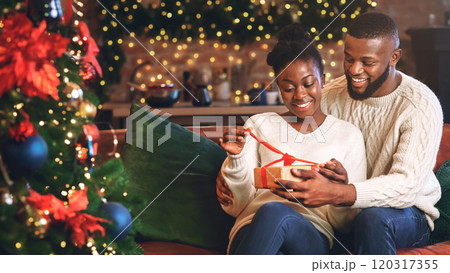 Happy african american woman unwrapping her christmas present with husband, celebrating holidays together at home. Happy african american woman unwrapping her christmas present with husband, celebrating holidays together at home. 120317355
