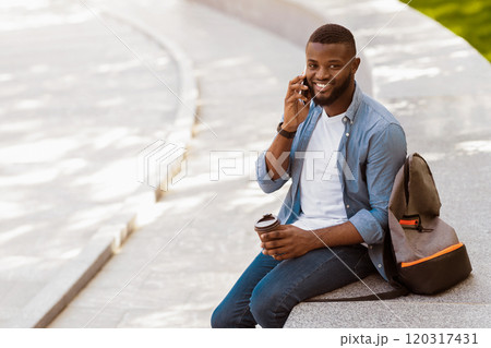 Handsome black man talking on phone outdoors, sitting on concrete bench, drinking takeaway coffee and looking at camera, copy space 120317431