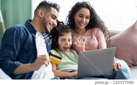 Cheerful Arabic Family Of Three With Laptop And Credit Card Making Online Shopping While Relaxing On Couch At Home, Happy Middle Eastern Parents And Little Daughter Enjoying Purchase From Internet 120317480
