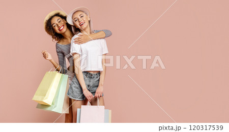 This image features two young women, one Black and one White, who are smiling and hugging each other. They are both dressed in casual summer clothing and are holding shopping bags. This image features two young women, one Black and one White, who are smiling and hugging each other. They are both dressed in casual summer clothing and are holding shopping bags. 120317539
