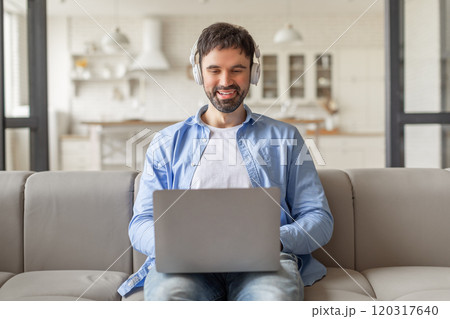 A man sits on a light gray couch in a modern living room, smiling while working on his laptop. He is wearing headphones and a light blue button-up shirt. 120317640