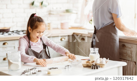 A young girl in a pink shirt and a grey apron is rolling dough on a white countertop in a kitchen. Her mom is out of focus in the background, preparing other ingredients for baking. A young girl in a pink shirt and a grey apron is rolling dough on a white countertop in a kitchen. Her mom is out of focus in the background, preparing other ingredients for baking. 120317642