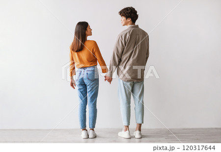 Full length of young Asian man and woman standing with their backs to camera, looking at each other and holding hands against white studio wall. Millennial couple expressing affection Full length of young Asian man and woman standing with their backs to camera, looking at each other and holding hands against white studio wall. Millennial couple expressing affection 120317644