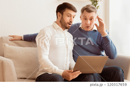 Oops, computer problem. Shocked elderly man and his adult son using laptop looking at error message sitting on sofa indoor 120317659