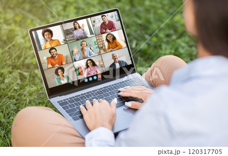 A person sits outside on a grassy lawn, using a laptop for a video conference. The screen displays a grid of nine people, each in a separate video frame A person sits outside on a grassy lawn, using a laptop for a video conference. The screen displays a grid of nine people, each in a separate video frame 120317705