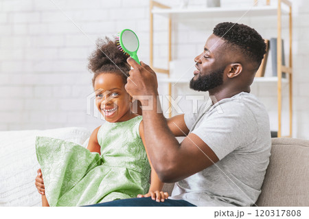 African American father is brushing his daughters hair while she sits on his lap. They are both smiling and looking happy. They are sitting on a couch in their home. 120317808